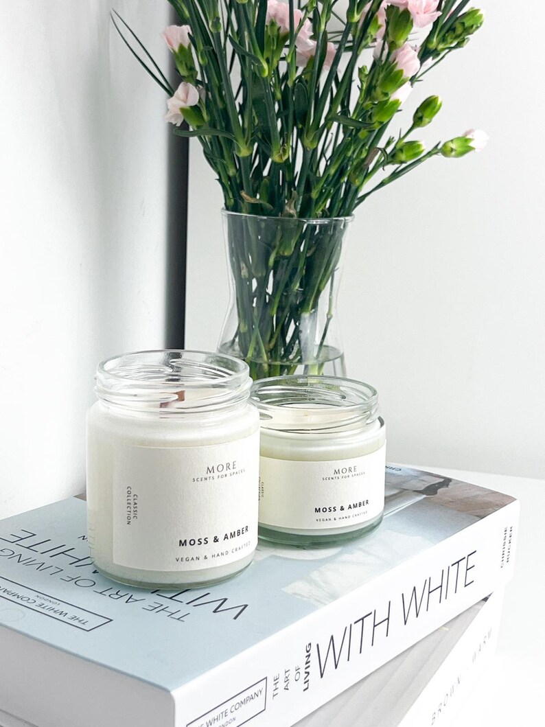 May include: Two white candle jars with black labels that read "More Scents for Spaces" and "Moss & Amber" on a stack of books with the text "The Art of Living With White" on the top book.