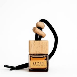 Peut inclure: Petite bouteille carr&eacute;e en verre avec un bouchon en bois et un cordon noir. La bouteille contient un liquide brun clair et porte l'inscription "MORE SCENTS FOR SPACES". Le bouchon en bois est en bois naturel.