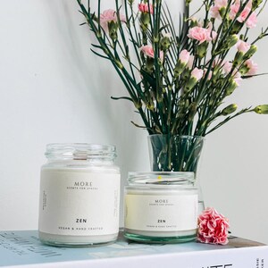 May include: Two glass candle jars with white labels that read "MORE SCENTS FOR SPACES ZEN VEGAN & HAND CRAFTED". The candles are sitting on a book titled "WITH WHITE" by Chrissie Rucker.