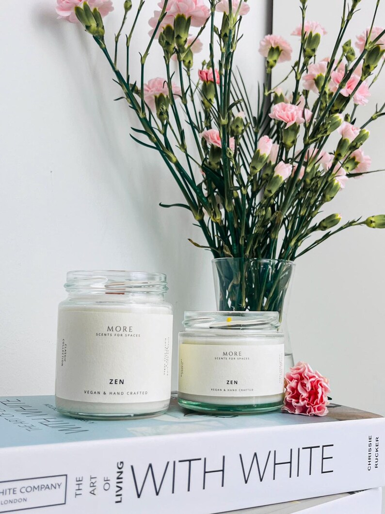 May include: Two glass candle jars with white labels that read "MORE SCENTS FOR SPACES ZEN VEGAN & HAND CRAFTED". The candles are sitting on a book titled "WITH WHITE" by Chrissie Rucker.