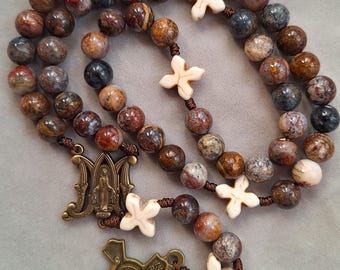 May include: A rosary featuring round, multi-colored brown and blue stone beads strung on brown cord. It includes white, cross-shaped spacer beads, a bronze-toned metal centerpiece depicting a figure, and a bronze-toned crucifix with INRI text.