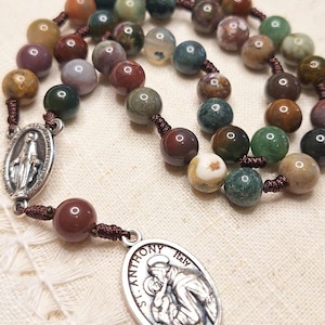 Puede incluir: Un rosario con cuentas redondas multicolores en tonos verdes, marrones, rojos y crema. Las cuentas están ensartadas en un cordón marrón. El rosario presenta dos colgantes plateados, uno con una imagen de la Virgen María y otro con el texto "ST. ANTHONY ITALY".