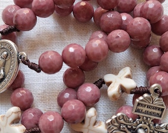 May include: A rosary with faceted, mauve-colored beads and off-white cross-shaped accents. The rosary features a silver crucifix and a medal with religious imagery. The beads are strung on a dark brown cord.