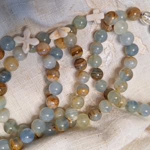 Peut inclure: Trois bracelets en perles bleues et brunes avec des breloques en forme de croix blanches. Les bracelets sont sur un fond en tissu blanc.
