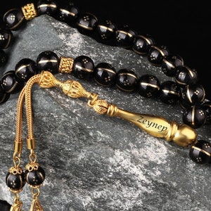May include: Black prayer beads with gold accents and a gold tassel. The tassel has the name "Zeynep" engraved on it.