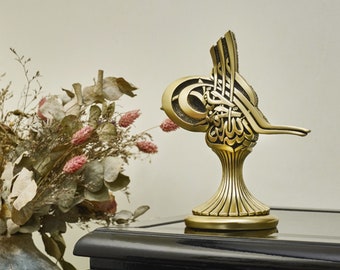 Pode incluir: Uma escultura de metal dourado de um pássaro estilizado com caligrafia intrincada em escrita árabe. A escultura está sobre uma superfície preta.