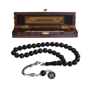 May include: Black prayer beads with a silver tassel and a silver charm inscribed with the words "Elif Semra" and an infinity symbol. The beads are presented in a wooden box with a carved design and a gold plaque featuring Turkish text.
