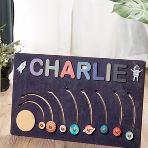 May include: A wooden board with the name "CHARLIE" spelled out in colorful letters. The board has a dark blue background with a rocket and astronaut design. There are eight planets with rings around them, and a sun at the top.