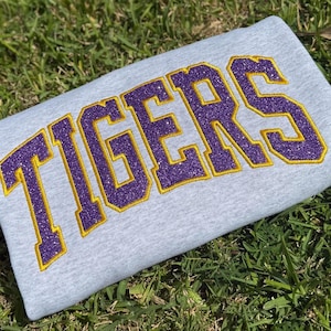 May include: A light gray sweatshirt with the word "TIGERS" embroidered in purple glitter with a gold outline. The sweatshirt is folded and resting on green grass.