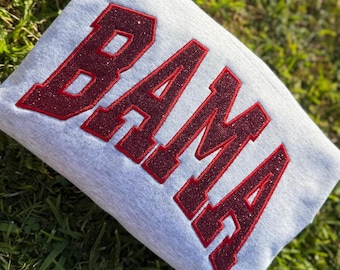 May include: A light gray sweatshirt with the word "BAMA" in large, glittery, maroon letters. The letters are outlined with a darker maroon color. The sweatshirt is folded and resting on a bed of green grass.