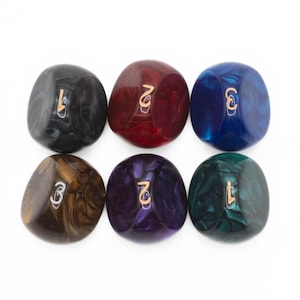 May include: Six colorful, marbled dice with gold-colored numbers. The dice are in shades of black, red, blue, brown, purple, and green. Each die has a different number from 1 to 7. The dice are smooth and rounded.