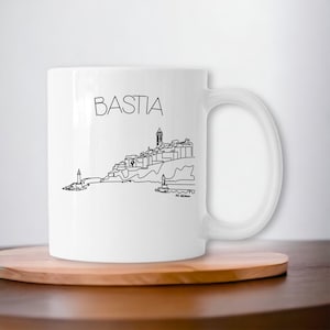Op de afbeelding: Witte keramische mok met een zwarte lijntekening van de stad Bastia, Corsica, Frankrijk. De mok heeft een zwart handvat en de tekst "BASTIA" is bovenaan in het zwart gedrukt.