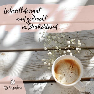 K&ouml;nnte beinhalten: Eine wei&szlig;e Keramiktasse mit Henkel, gef&uuml;llt mit Kaffee. Der Kaffee hat eine hellbraune Farbe und einen schaumigen Belag. Die Tasse steht auf einer wei&szlig;en Holzoberfl&auml;che mit einem Zweig wei&szlig;er Bl&uuml;ten. Der Text "Liebevoll designt und gedruckt in Deutschland" ist in Schwarz auf rosa Hintergrund geschrieben.