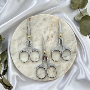May include: Three silver embroidery scissors with ornate handles and gold-colored rivets are arranged on a marble-like circular surface. The scissors are set against a backdrop of white satin fabric and greenery.