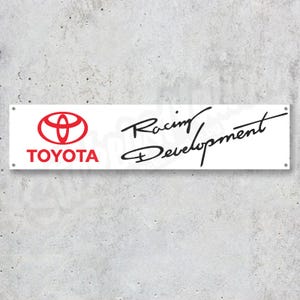 Op de afbeelding: Rechthoekige witte banner met het Toyota-logo in rood en het woord "TOYOTA" in rood. De woorden "Racing Development" zijn in zwart script geschreven. De banner heeft grommets in elke hoek.