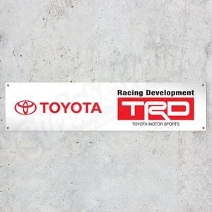 Könnte beinhalten: Weißes rechteckiges Schild mit dem Toyota-Logo in Rot auf der linken Seite und dem TRD Racing Development-Logo auf der rechten Seite. Das Schild enthält auch den Text "TOYOTA MOTOR SPORTS".