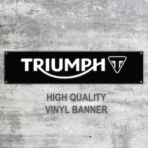 Könnte beinhalten: Ein schwarzes rechteckiges Vinylbanner mit dem weißen Wort "TRIUMPH" und einem Logo. Das Banner hat auch den Text "HIGH QUALITY VINYL BANNER" in Grau. Das Banner ist an einer grauen Betonwand befestigt.