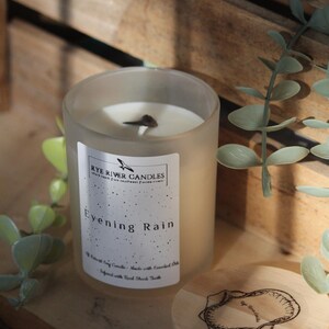 May include: A frosted glass candle jar with a white label that reads "Rye River Candles" and "Evening Rain". The candle is on a wooden surface with green leaves.