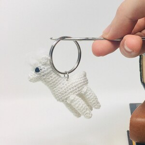 Op de afbeelding: Een witte gehaakte alpaca sleutelhanger met een zilveren sleutelring. De alpaca heeft een pluizige kop en kleine blauwe ogen. De sleutelhanger wordt omhoog gehouden door een zilveren staaf.