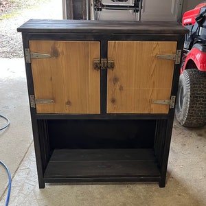 May include: A black wooden cabinet with two doors and a shelf. The doors are made of light wood and have metal hinges and latches. The cabinet is standing on a light-colored floor.