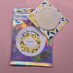 May include: A holographic pouch containing scented putty with a circular label reading It's So Rachel Scented Putty Lemon featuring a lemon wreath illustration. A square card with a lemon pattern sits behind a round, off-white putty disc.