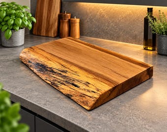 Puede incluir: Una tabla de cortar rectangular de madera con un borde natural en uno de sus lados. La tabla muestra una veta de madera de color marrón claro cálido con detalles de corteza oscura y texturizada a lo largo del borde irregular.