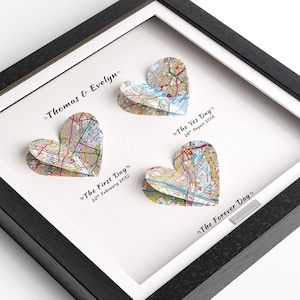 Puede incluir: Una obra de arte enmarcada con tres recortes de mapas en forma de corazón. La obra de arte incluye los nombres "Thomas & Evelyn" y las fechas: "The First Day 22 de febrero de 2012", "The Yes Day 28 de agosto de 2016" y "The Forever Day". El marco es negro.