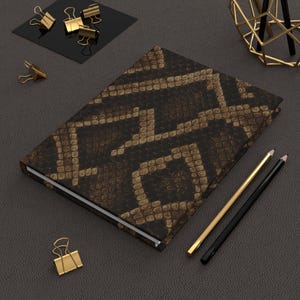 May include: A hardcover journal with a snakeskin pattern in shades of brown and tan. Gold-colored binder clips and pencils are arranged around the journal on a dark gray surface. The journal is closed.