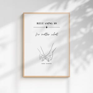 May include: A framed wall art print featuring a minimalist black line drawing of two hands holding each other. The text reads RELEASING 10, no matter what, and LIZZIE AND HUGH. The design is centered on a white background within a light wood frame.