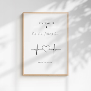 May include: A rectangular wall art print in a light wood frame featuring black text on a white background. The text reads BINDING 13, Boom boom fucking boom, and JOHNNY AND SHANON. A black heartbeat line graphic with a central heart shape is included.