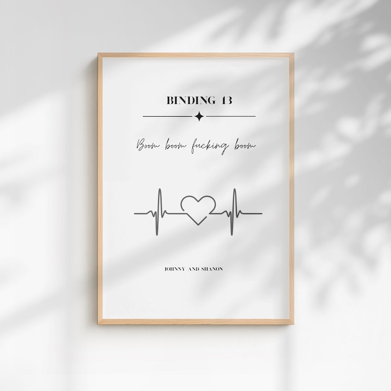 May include: A rectangular wall art print in a light wood frame featuring black text on a white background. The text reads BINDING 13, Boom boom fucking boom, and JOHNNY AND SHANON. A black heartbeat line graphic with a central heart shape is included.