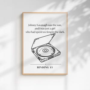 May include: A white rectangular poster in a light wood frame featuring black line art of an open CD player with a disc inside labeled SHANNON'S SONGS FOR JOHNNY. The text reads: Johnny Kavanagh was the sun, and I was just a girl who had spent too long in the dark. BINDING 13.