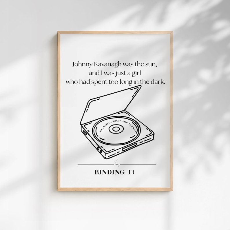 May include: A white rectangular poster in a light wood frame featuring black line art of an open CD player with a disc inside labeled SHANNON'S SONGS FOR JOHNNY. The text reads: Johnny Kavanagh was the sun, and I was just a girl who had spent too long in the dark. BINDING 13.