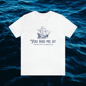 Può includere: Maglietta bianca con un'illustrazione blu di una nave e il testo "You had me at trigger warning."