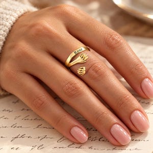 May include: A gold-toned open ring featuring two hands meeting in the center, with the name Rose engraved on the band. The design wraps around the finger, creating a symbolic embrace with detailed fingers and palms on the metal surface.