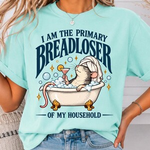 May include: A light mint green t-shirt featuring a graphic of a rat in a clawfoot bathtub filled with bubbles, wearing a towel on its head and holding a cocktail. The text reads I AM THE PRIMARY BREADLOSER OF MY HOUSEHOLD in dark blue lettering.