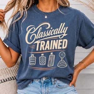 May include: A blue short-sleeve t-shirt featuring the cream-colored text Classically Trained above a graphic illustration of three car pedals and a manual gear shifter. The design is centered on the chest of the garment.