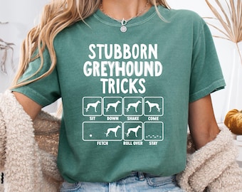 May include: A teal green t-shirt with the text "Stubborn Greyhound Tricks" in white. Below the text are illustrations of a greyhound demonstrating various tricks, including "Sit", "Down", "Shake", "Come", "Fetch", "Roll Over", and "Stay".