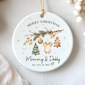 May include: A round white ceramic Christmas ornament hanging from a gold string. The design features a pine branch with baby-themed decorations, including a bottle, socks, a onesie, and trees. Text reads: MERRY CHRISTMAS Mommy & Daddy SEE YOU IN 2027.