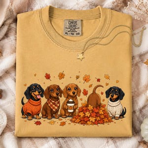 May include: A mustard yellow t-shirt featuring a graphic of five dachshunds in autumn attire, with some wearing sweaters and scarves, one holding a coffee cup, and one playing in a pile of fall leaves. The shirt has a Comfort Colors brand label.