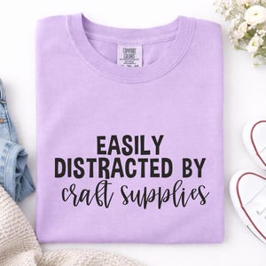 May include: A lavender Comfort Colors crewneck sweatshirt with the text "EASILY DISTRACTED BY craft supplies" in black. The shirt is folded neatly, with a pair of white sneakers and a sprig of white flowers visible in the background.