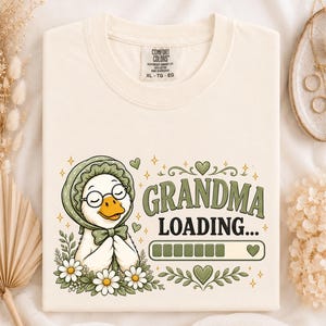 May include: A cream-colored t-shirt featuring a graphic of a white goose wearing a green bonnet, glasses, and a bow tie. The goose is surrounded by white daisies. The text reads "GRANDMA LOADING..." above a green progress bar and a heart icon.