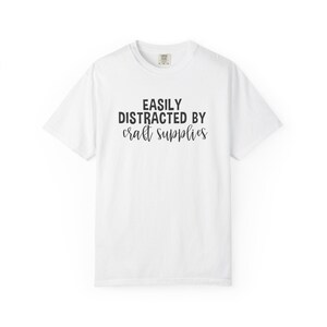 May include: White t-shirt with the text "EASILY DISTRACTED BY craft supplies" in black. The shirt has a crew neck and short sleeves. The text is in a bold, sans-serif font.