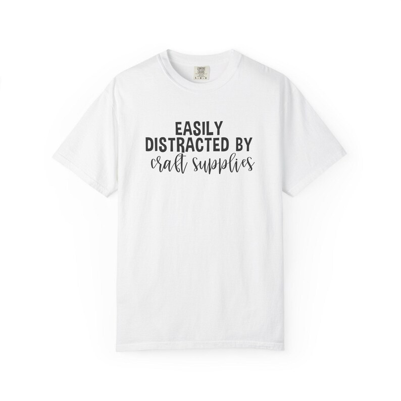 May include: White t-shirt with the text "EASILY DISTRACTED BY craft supplies" in black. The shirt has a crew neck and short sleeves. The text is in a bold, sans-serif font.