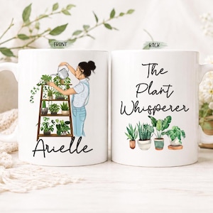 May include: Two white ceramic mugs featuring illustrations. The front mug shows a person watering plants on a wooden shelf with the name Arielle below. The back mug displays the text The Plant Whisperer above a collection of four potted plants.