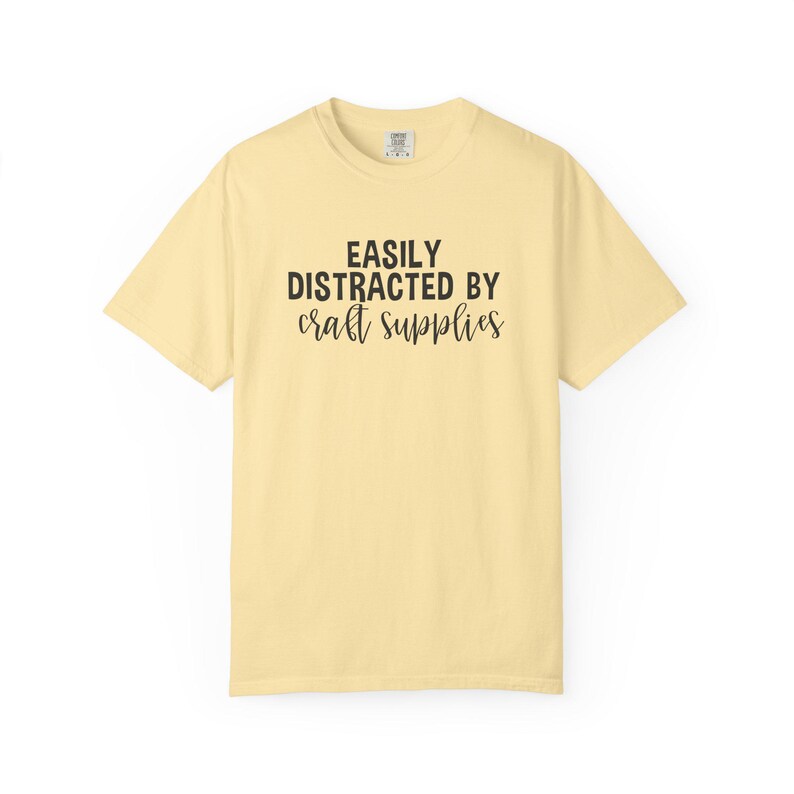 May include: A light yellow t-shirt with the text "EASILY DISTRACTED BY craft supplies" in black. The shirt has short sleeves and a crew neck. The shirt is made of a soft, comfortable material.