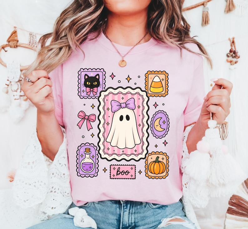 May include: A light pink t-shirt featuring a central graphic of a white ghost wearing a purple bow. Surrounding the ghost are smaller framed illustrations of a black cat, candy corn, a crescent moon, a pumpkin, and a potion bottle with the text boo.