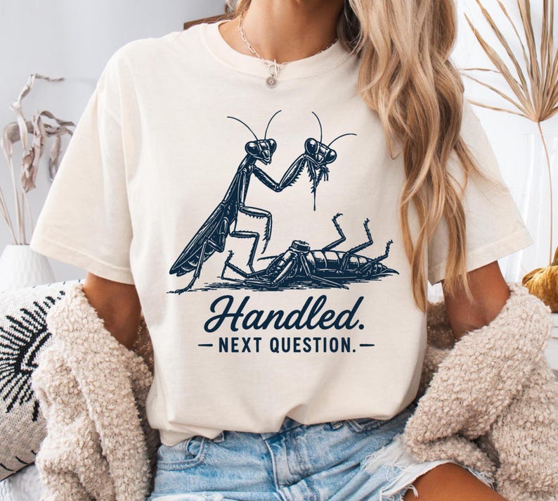 May include: A cream-colored short-sleeve t-shirt featuring a dark blue graphic illustration of a praying mantis standing over a cockroach. Below the illustration, the text reads "Handled. - NEXT QUESTION. -" in a dark blue, bold, serif font.