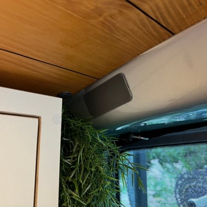 Puede incluir: Un armario blanco con un borde de madera y una pequeña planta colgando de la parte superior. El armario está en una furgoneta con un techo gris y una cortina de ventana de malla negra.