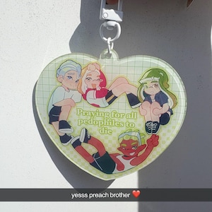 Puede incluir: Llavero en forma de corazón con una ilustración de dibujos animados de cuatro figuras. El texto dice: "Praying for all pedophiles to die." El llavero tiene un cierre blanco.